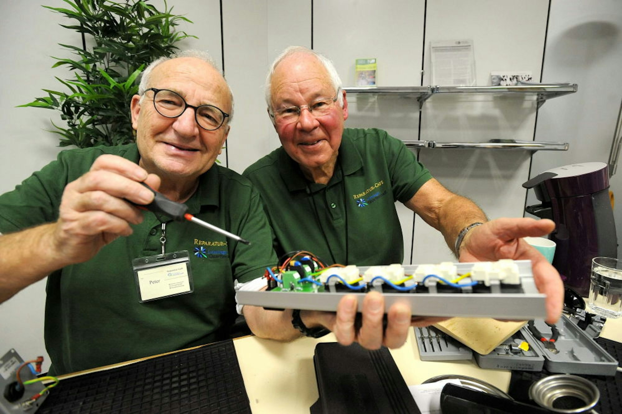 Die Ehrenamtler Peter Esch (l.) und Martin Sylla, in grünen Polo-Shirts des Helferteams, mit einer defekten Steckdosenleiste.