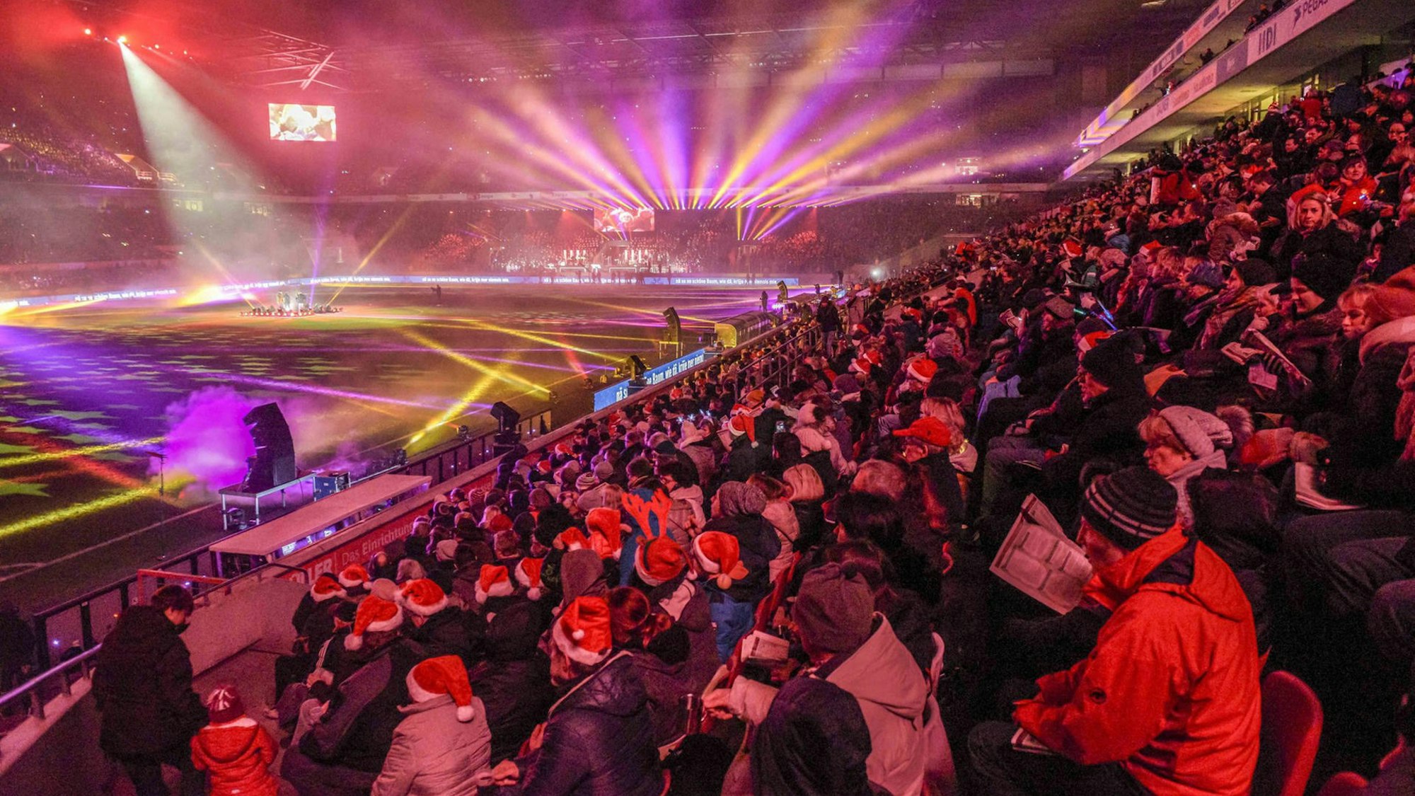 Von der Tribüne des Rhein-Energie-Stadion schauen Tausende Menschen, viele mit Weihnachtsmannmützen auf dem Kopf, auf eine bunt erleuchtete Stadionfläche.