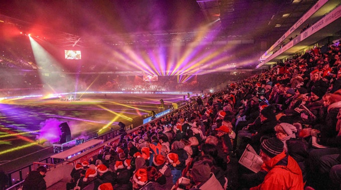 Von der Tribüne des Rhein-Energie-Stadion schauen Tausende Menschen, viele mit Weihnachtsmannmützen auf dem Kopf, auf eine bunt erleuchtete Stadionfläche.