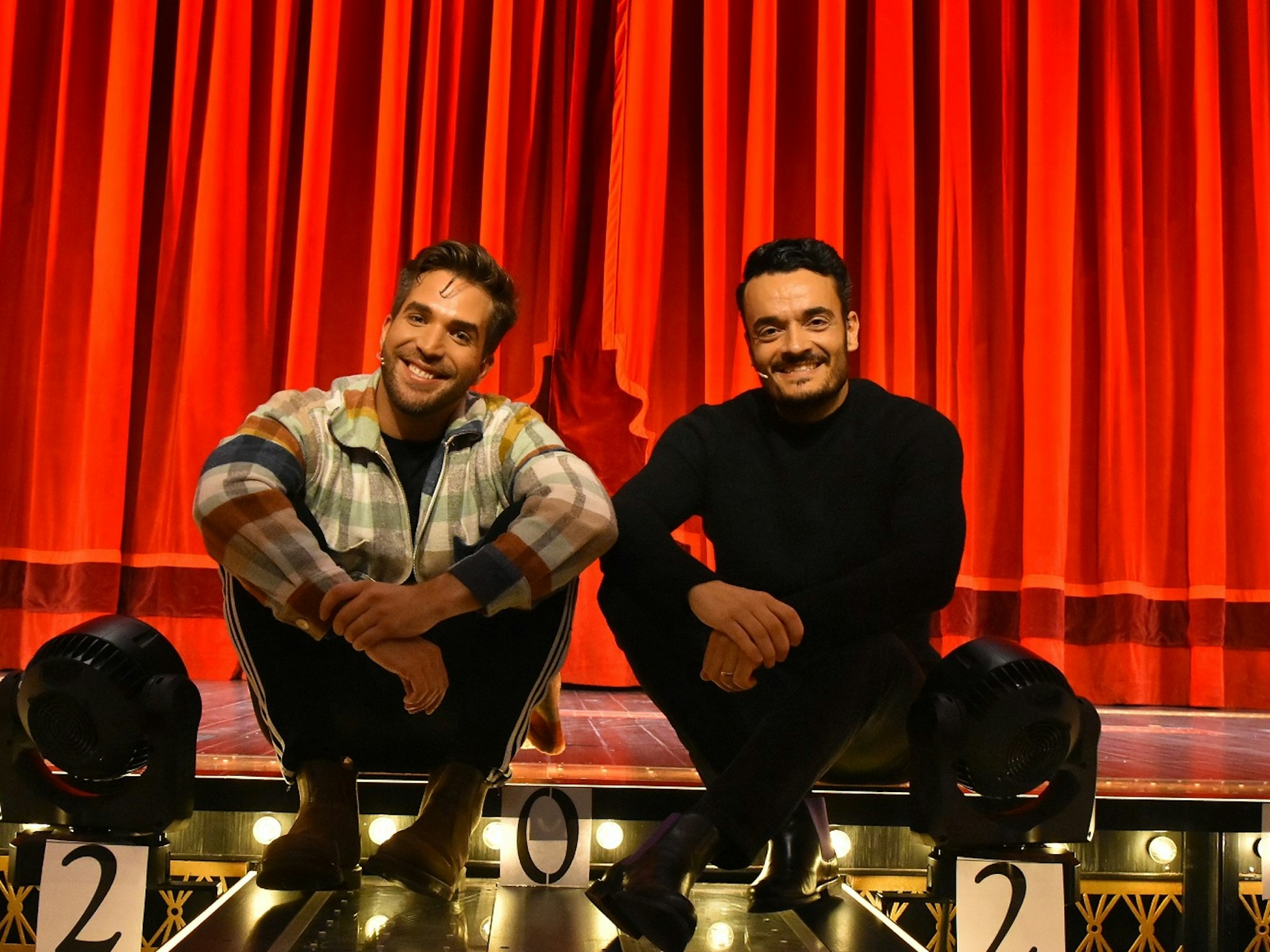 Riccardo Greco und Giovanni Zarrella sitzen lachend vor dem roten Vorhang auf der Bühne von Moulin Rouge.
