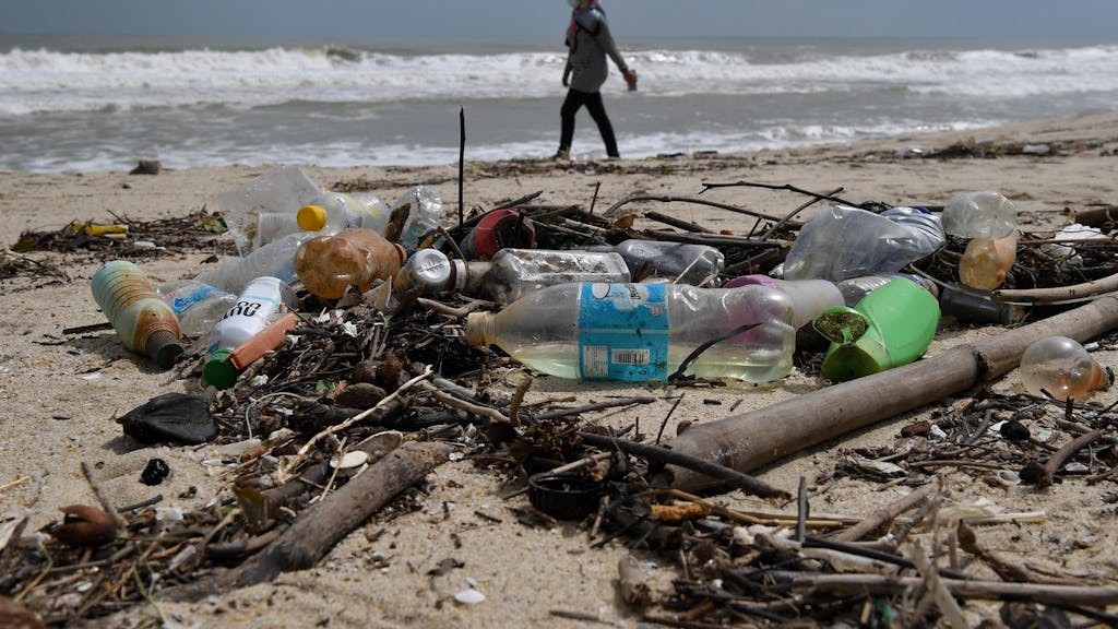 Frau geht am Strand spazieren, im Vordergrund liegen Plastikflaschen.
