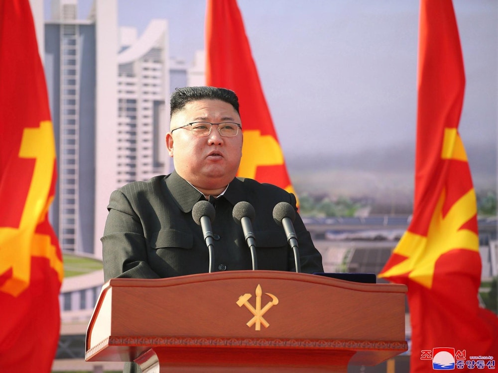 Auf dem Symbolfoto von 2021 steht Kim Jong Un hinter einer Holzkanzel mit drei Mikrofonen, im Hintergrund befinden sich drei Flaggen und Hochhäuser.