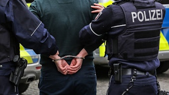 Zwei Polizisten führen einen Mann ab, dessen Hände auf dem Rücken gefesselt sind.
