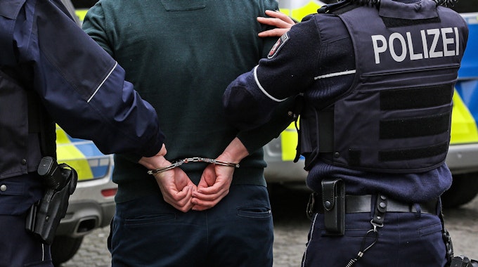 Zwei Polizisten führen einen Mann ab, dessen Hände auf dem Rücken gefesselt sind.