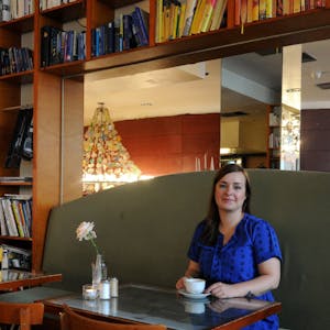 Julia Floß sitzt in einem blauen Kleid auf einem Sofa, vor ihr steht eine Tasse Kaffee, im Hintergrund ist ein Bücherregal zu sehen