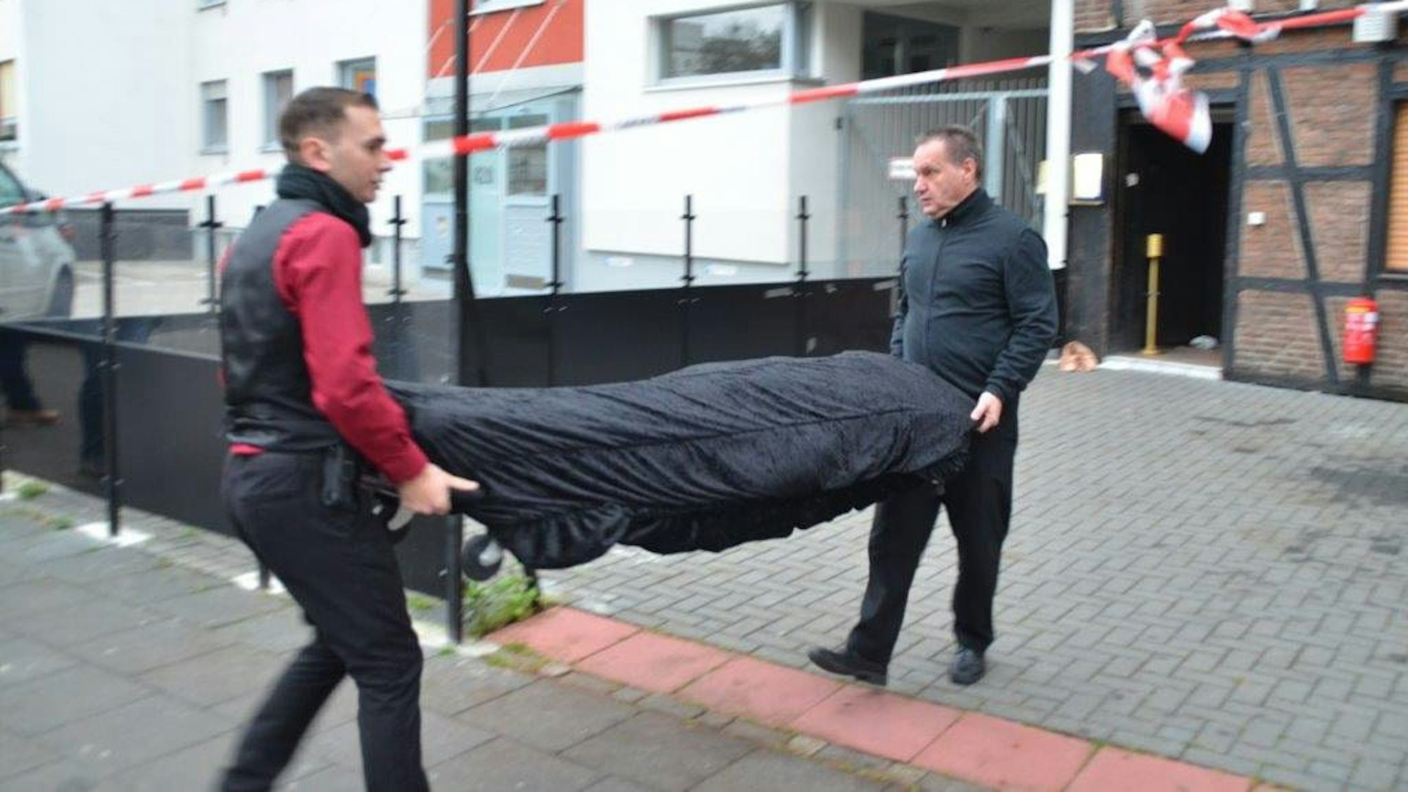 Vor einer Kneipe, die mit Flatterband der Polizei abgesperrt ist, tragen zwei Bestatter auf einer Bahre eine abgedeckte Leiche weg.
