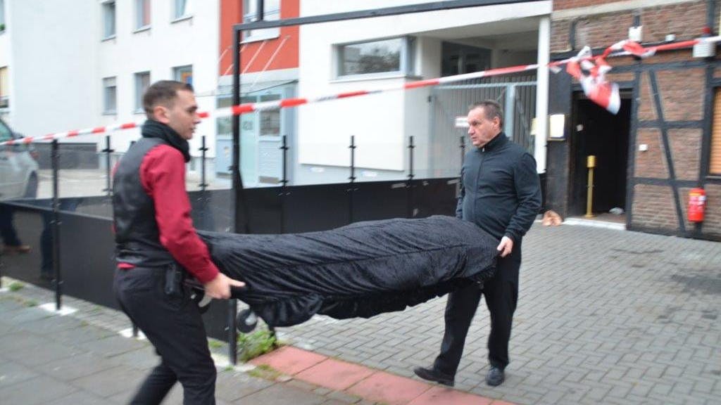 Vor einer Kneipe, die mit Flatterband der Polizei abgesperrt ist, tragen zwei Bestatter auf einer Bahre eine abgedeckte Leiche weg.&nbsp;
