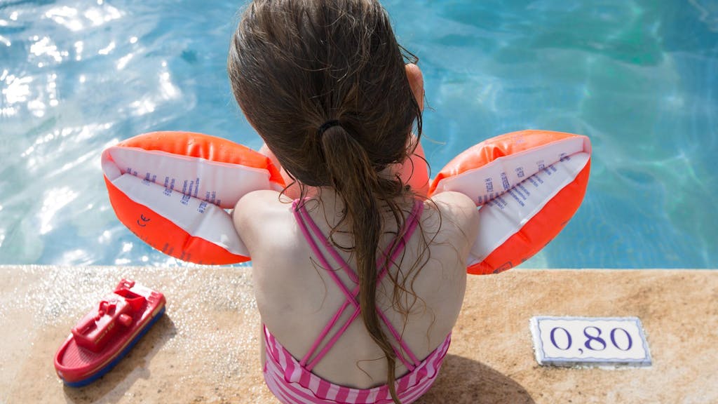 Ein Mädchen sitzt mit Schwimmflügeln am Beckenrand.