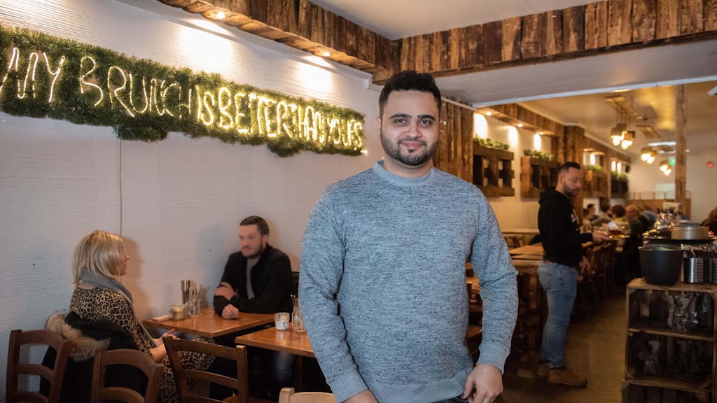 Parham Pooramin steht mit einem grauen Pullover im Café Buur in Köln, im Hintergrund sind Gäste zu sehen.