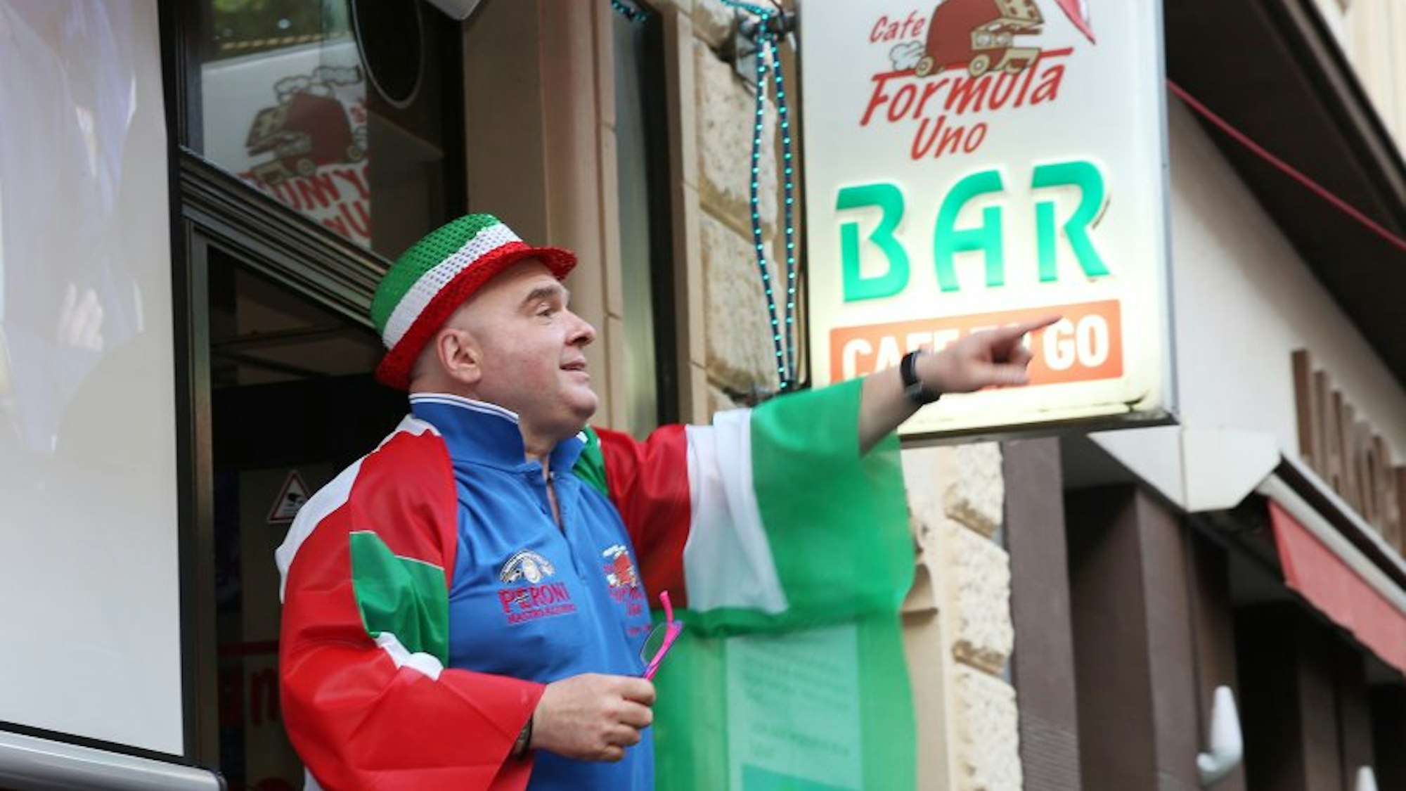 Mann mit Hut in italienischen Farben und Italien-Flagge umgehängt zeigt auf einen Punkt in der Ferne