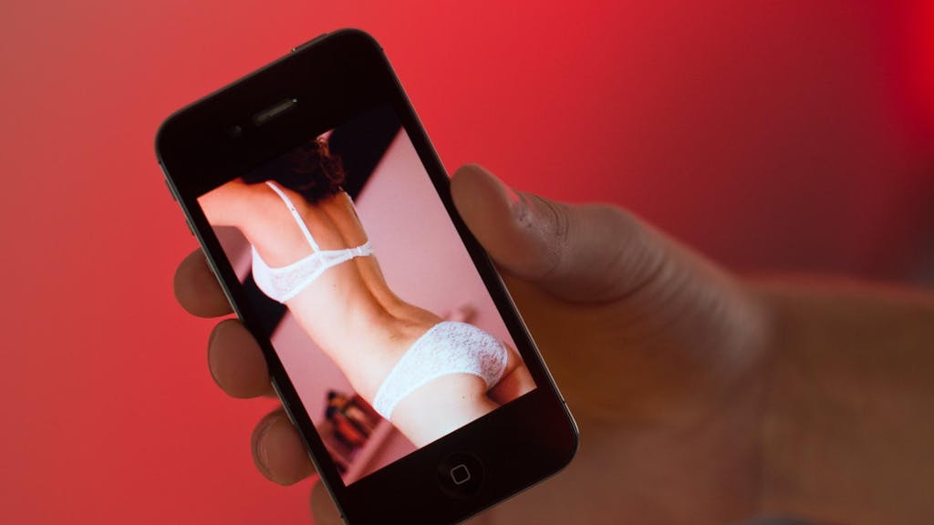 Ein junger Mann hält ein Smartphone, auf dem ein erotisches Foto einer jungen erwachsenen Frau zu sehen ist.