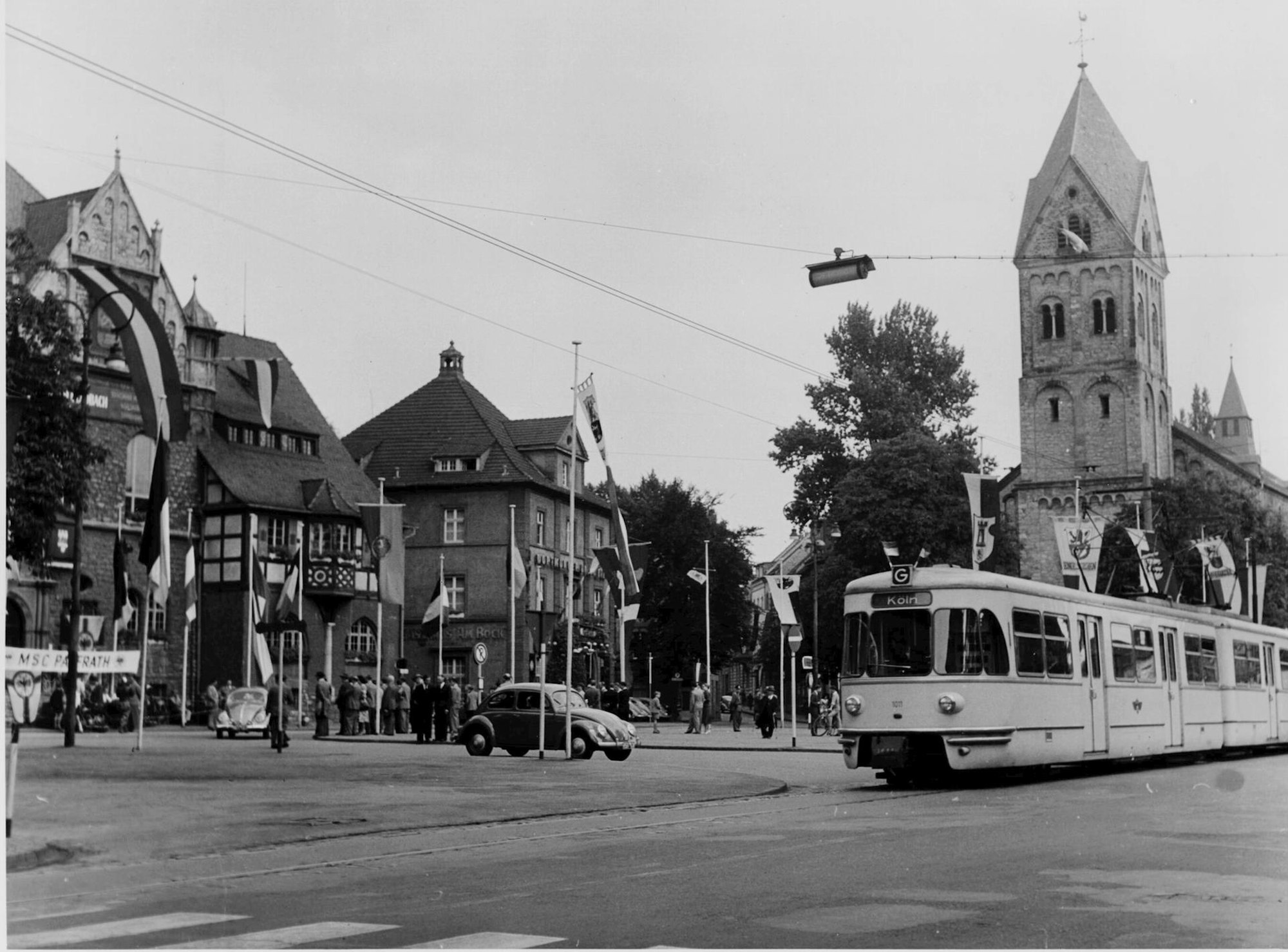 Blick auf den fahnengeschmückten Marktplatz in Bergisch Gladbach während der Jahrhundertfeier am 22. Juli 1956. Die Linie G fährt an diesem Tag in Richtung Köln. Rechts im Hintergrund ist die Kirche St. Laurentius zu sehen.