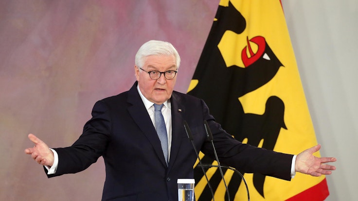 Das Symbolfoto von 2021 zeigt den deutschen Bundespräsidenten Frank-Walter Steinmeier. Er steht an einem Rednerpult mit drei Mikrofonen, hinter ihm befindet sich eine Flagge.