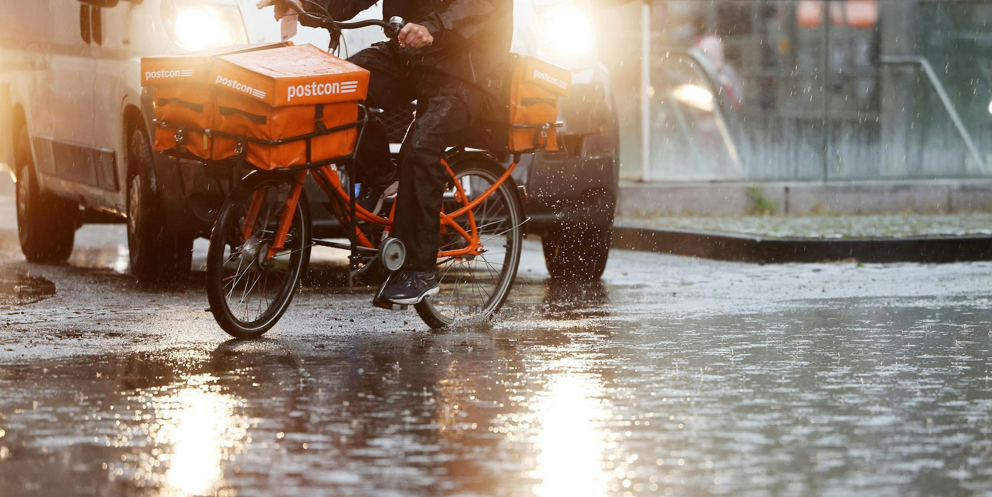 Ein Postbote fährt mit seinem Fahrrad über eine regennasse Straße. Im Hintergrund ist ein Transporter mit Scheinwerfern zu sehen. (Symbolbild)