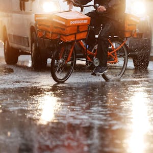Ein Postbote fährt mit seinem Fahrrad über eine regennasse Straße. Im Hintergrund ist ein Transporter mit Scheinwerfern zu sehen. (Symbolbild)