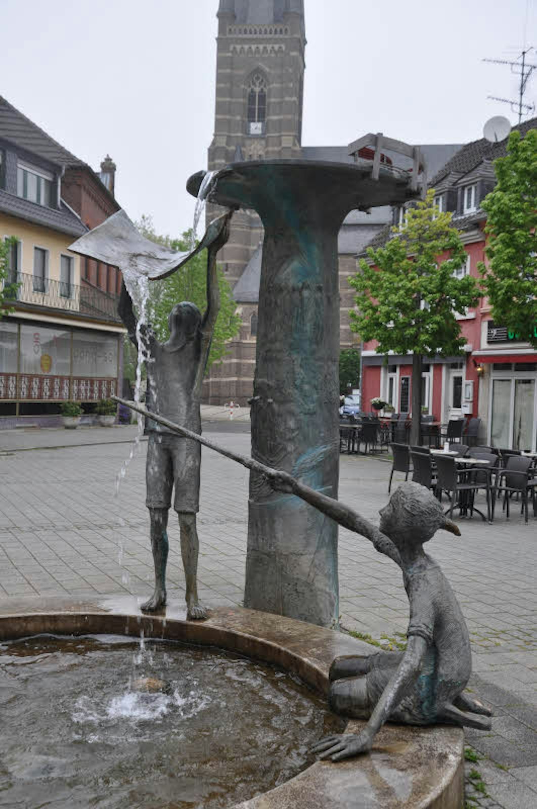 Auf dem Bedburger Marktplatz plätschert das Wasser aus einem originell gestalteten Brunnen des Künstlers Juan Fernandez, der den Titel „Geschichte, Kinder und Wasser“ trägt.