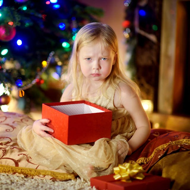 Ein Kind hält ein Weihnachtsgeschenk und sitzt vor dem Tannenbaum, es schaut schlecht gelaunt.