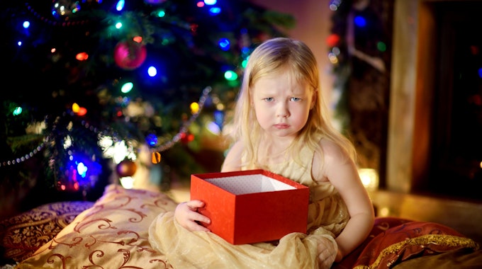 Ein Kind hält ein Weihnachtsgeschenk und sitzt vor dem Tannenbaum, es schaut schlecht gelaunt.