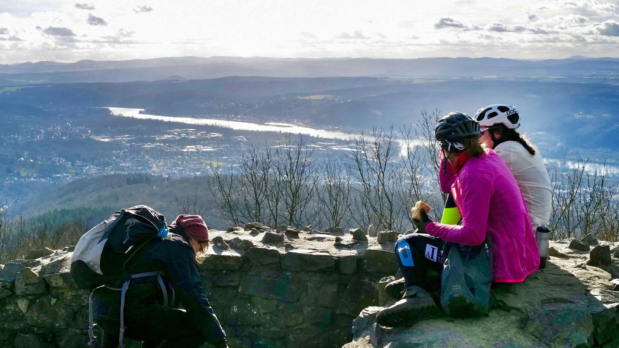 Drei Frauen machen auf dem Gipfel eines Berges Pause und sitzen dabei auf einer Steinmauer.