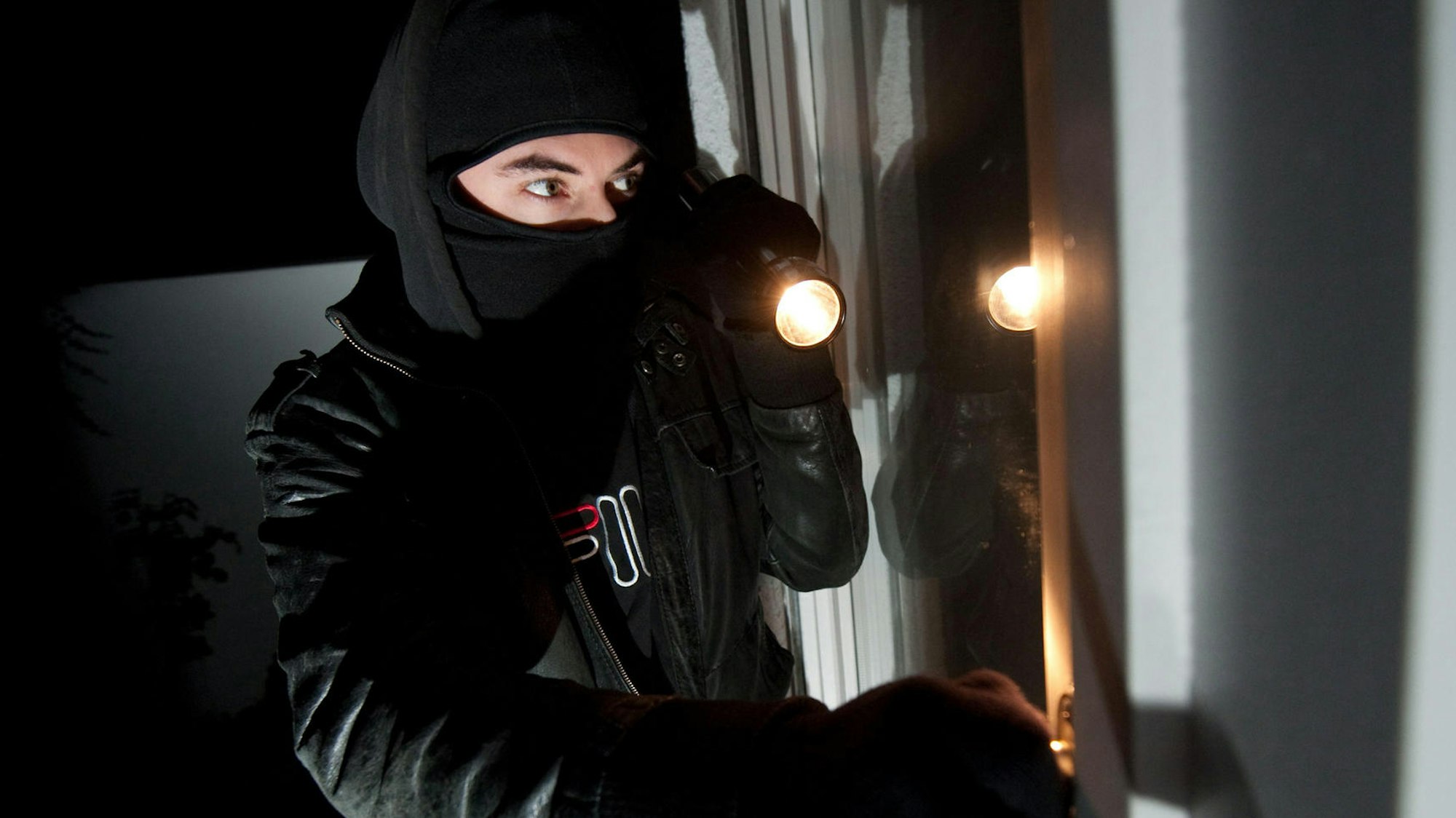 Ein vermummter Einbrecher leuchtet mit einer Taschenlampe in eine Wohnung.