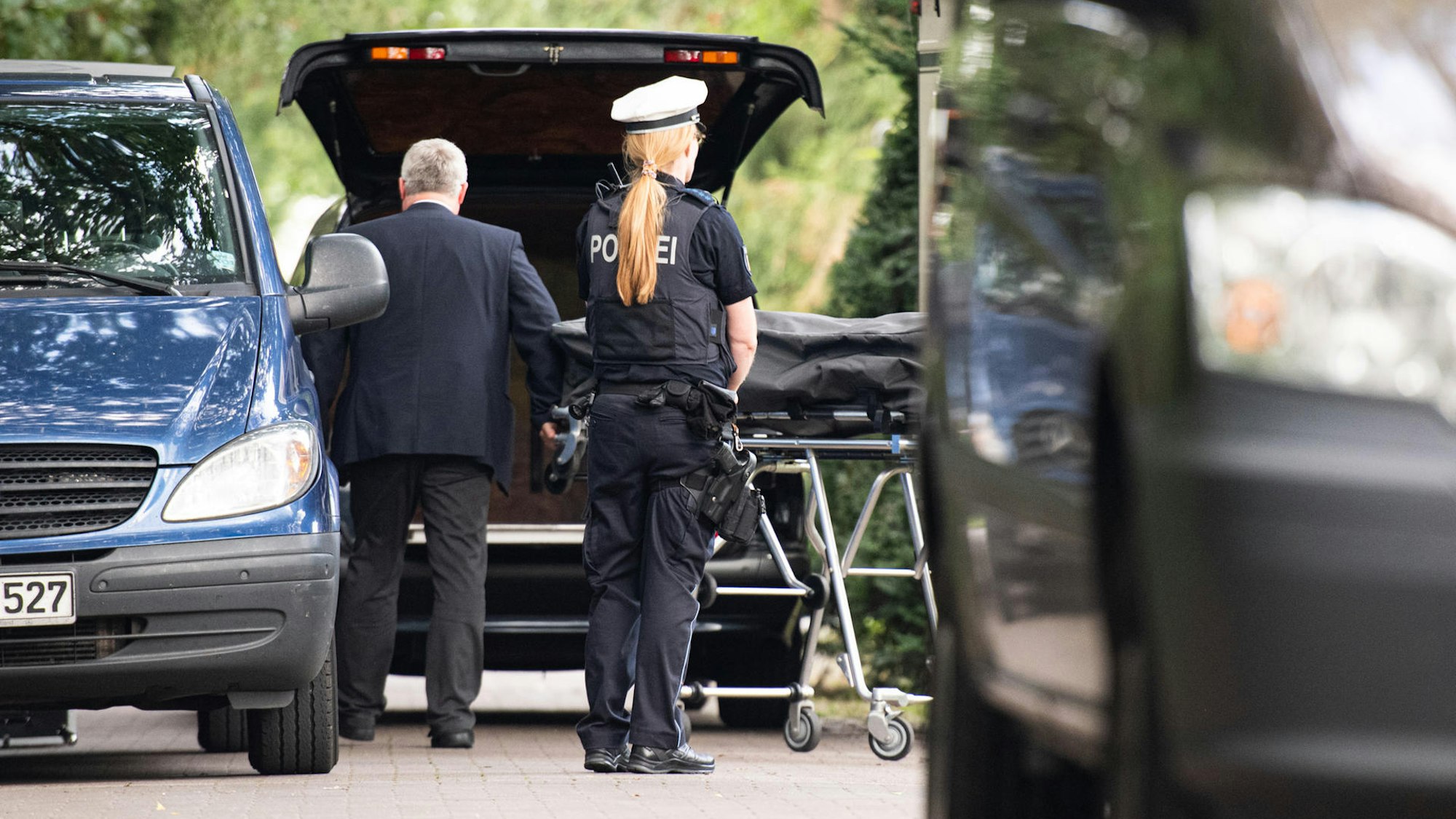Ein Bestatter lädt einen Leichnam, der sich in einem Leichensack befindet, in sein Auto. Davor steht eine Polizistin.