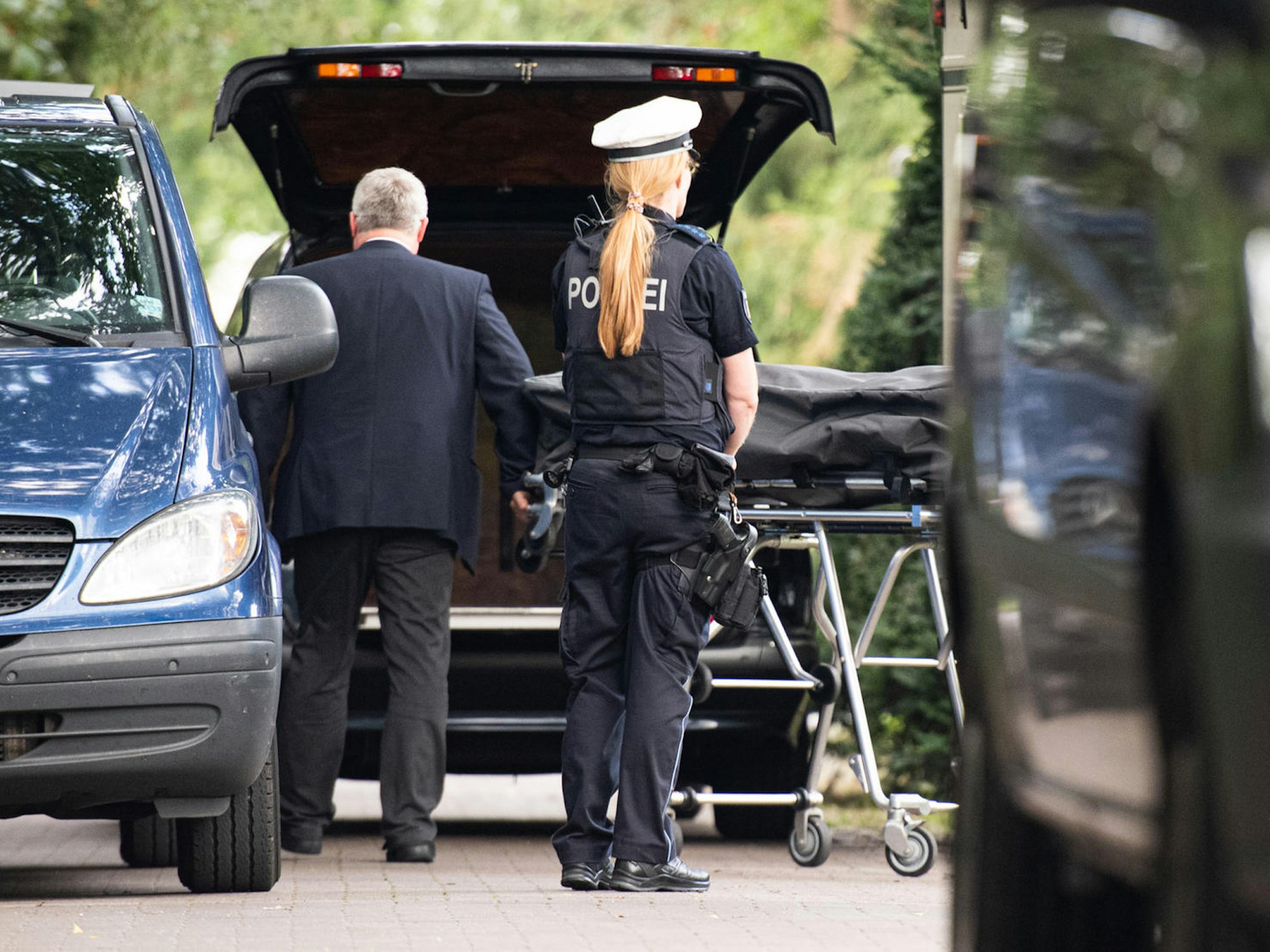 Ein Bestatter bringt einen Leichnam zu seinem Wagen, davor steht eine Polizistin.