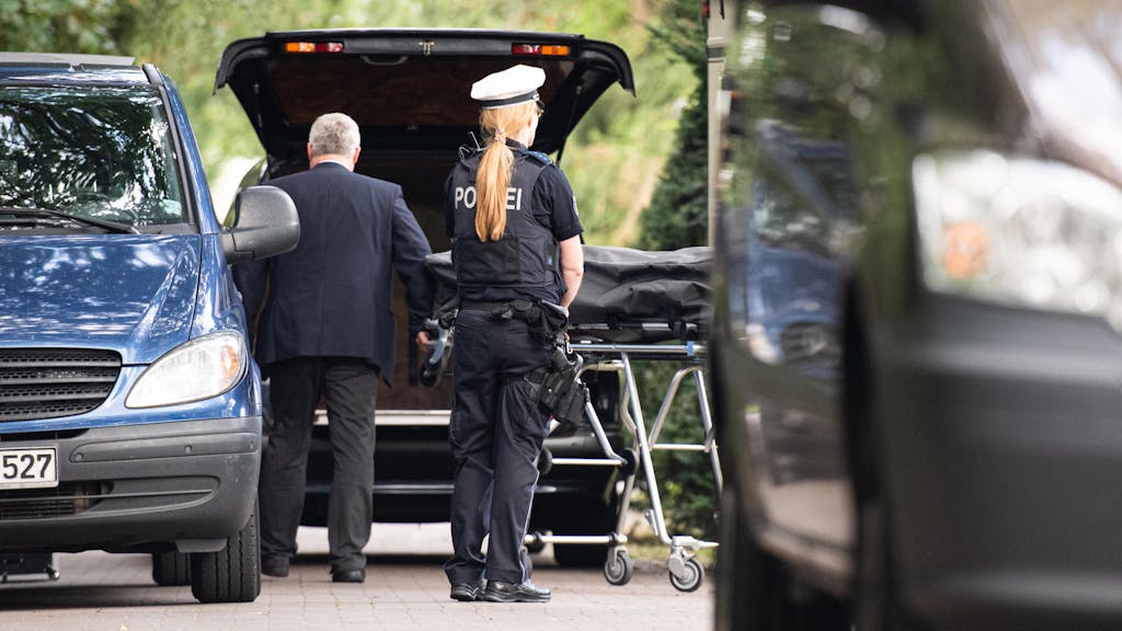 Ein Bestatter lädt einen Leichnam, der sich in einem Leichensack befindet, in sein Auto. Davor steht eine Polizistin.&nbsp;