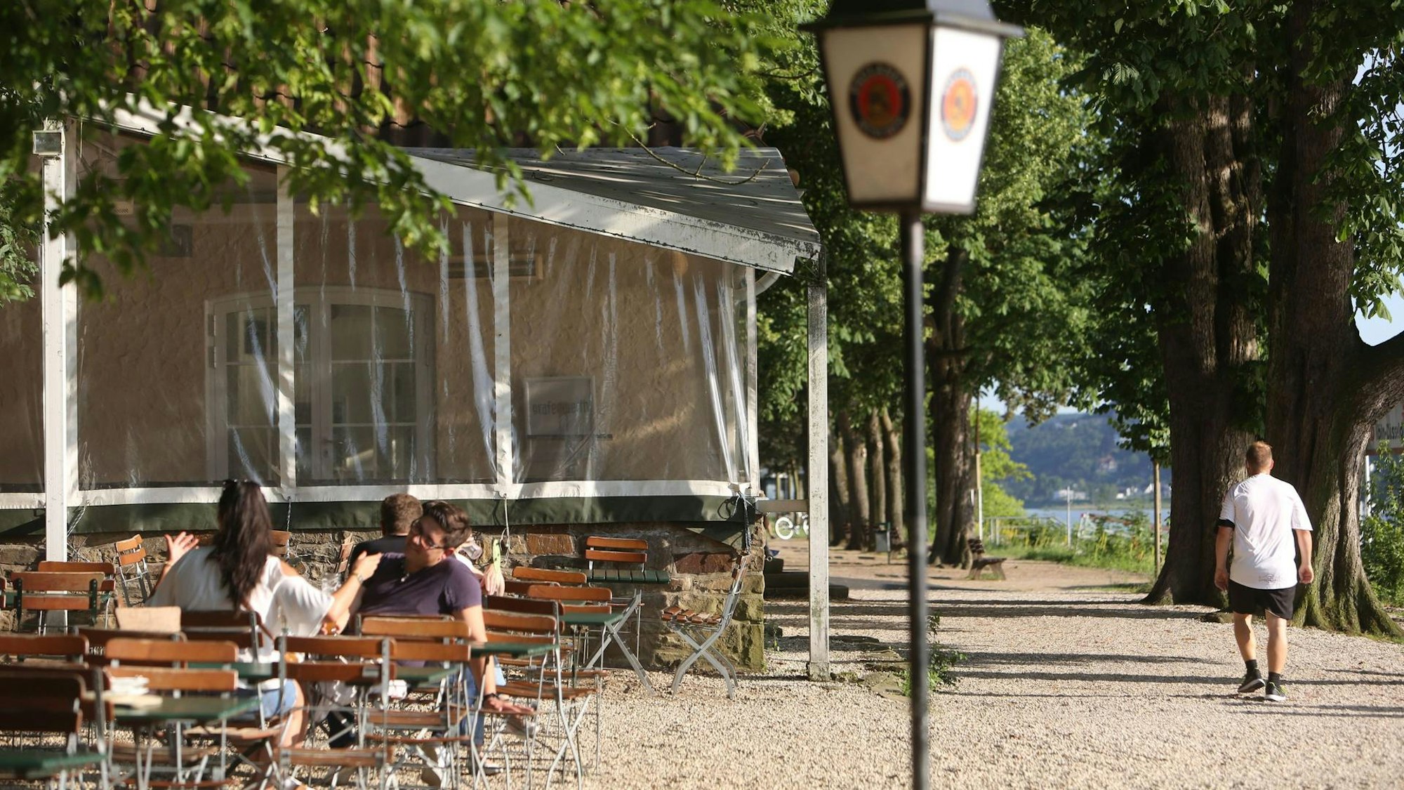 Tische und Stühle stehen unter Bäumen neben einem Fußweg, der direkt am Rhein liegt.
