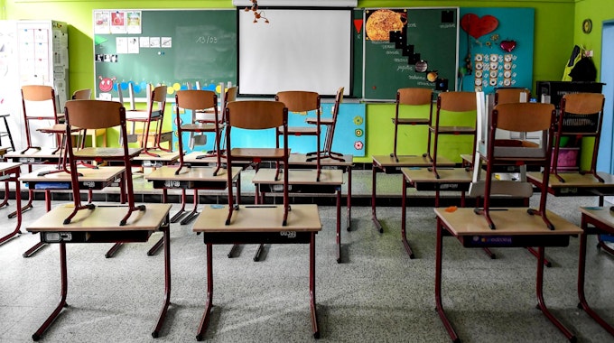 In einem leeren Klassenzimmer stehen Stühle auf Tischen. Vorne ist eine Tafel zu sehen, vor der Tafel ist eine Leinwand.