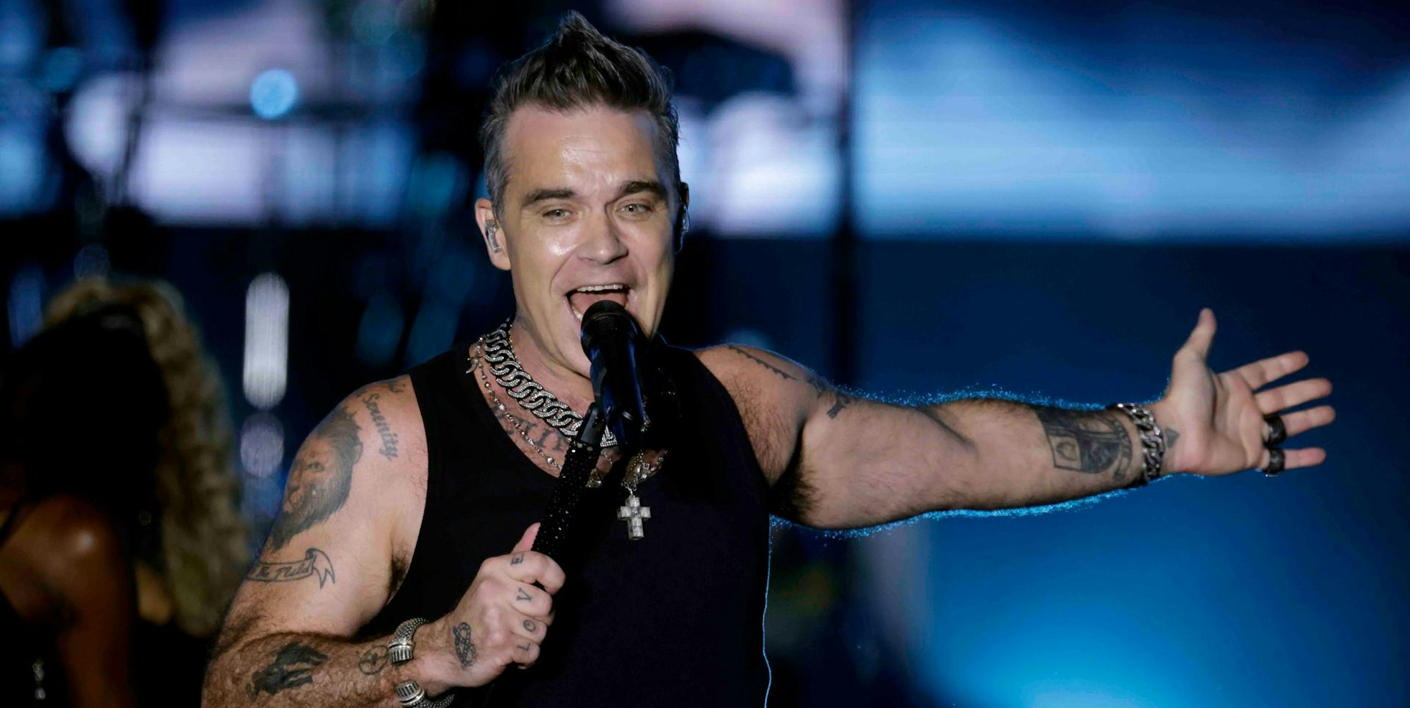 Der britische Sänger Robbie Williams am 30. August 2022 auf der Bühne im Bonner Hofgarten. Er singt ins Standmikrofon, trägt ein Muscle-Shirt und hat einen Arm ausgestreckt.
