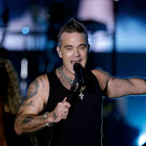 Der britische Sänger Robbie Williams am 30. August 2022 auf der Bühne im Bonner Hofgarten. Er singt ins Standmikrofon, trägt ein Muscle-Shirt und hat einen Arm ausgestreckt.