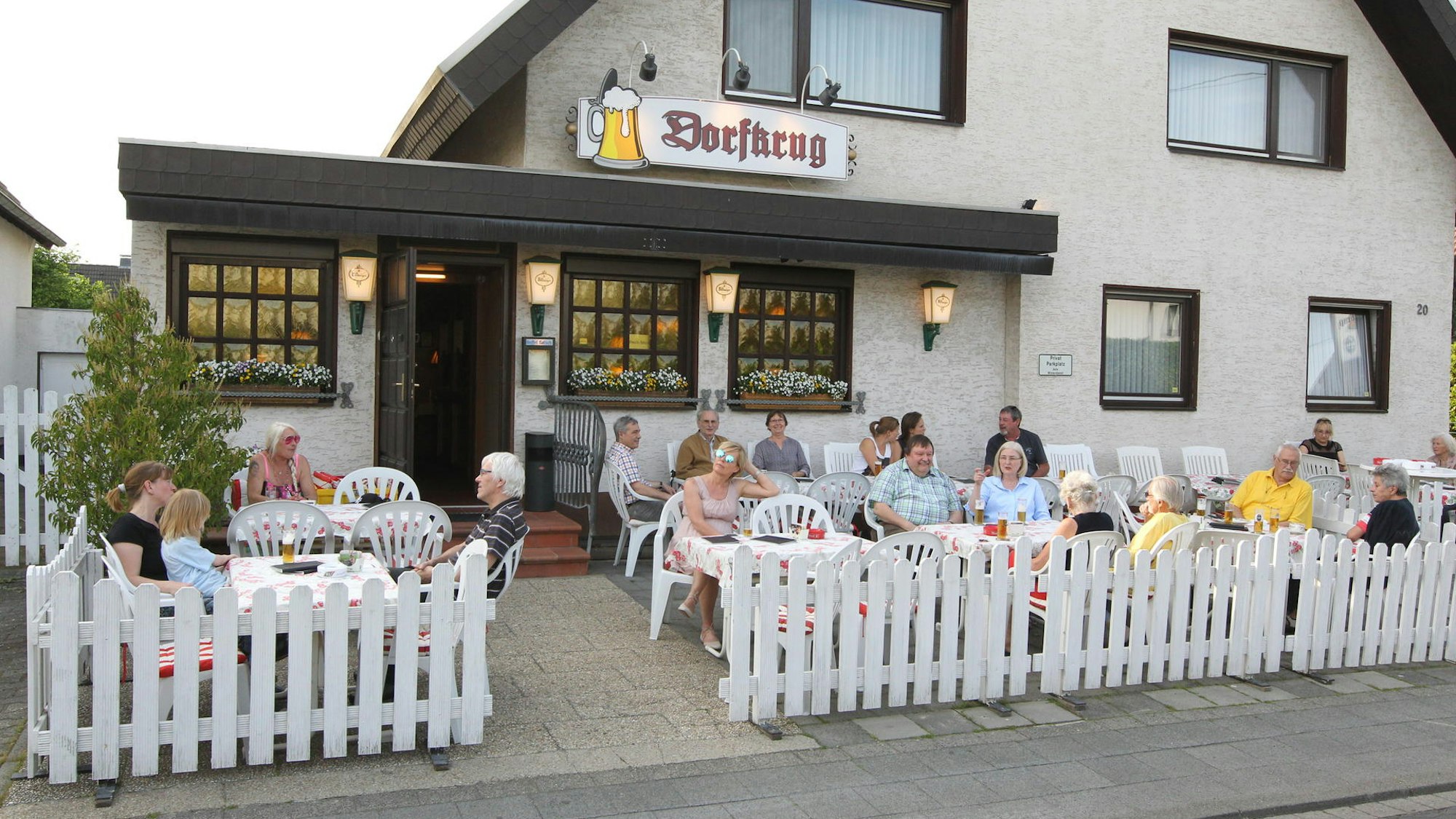 Vor einem Gebäude mit dem Schild „Dorfkurg“ sitzen mehrere Personen an Tischen im Freien.