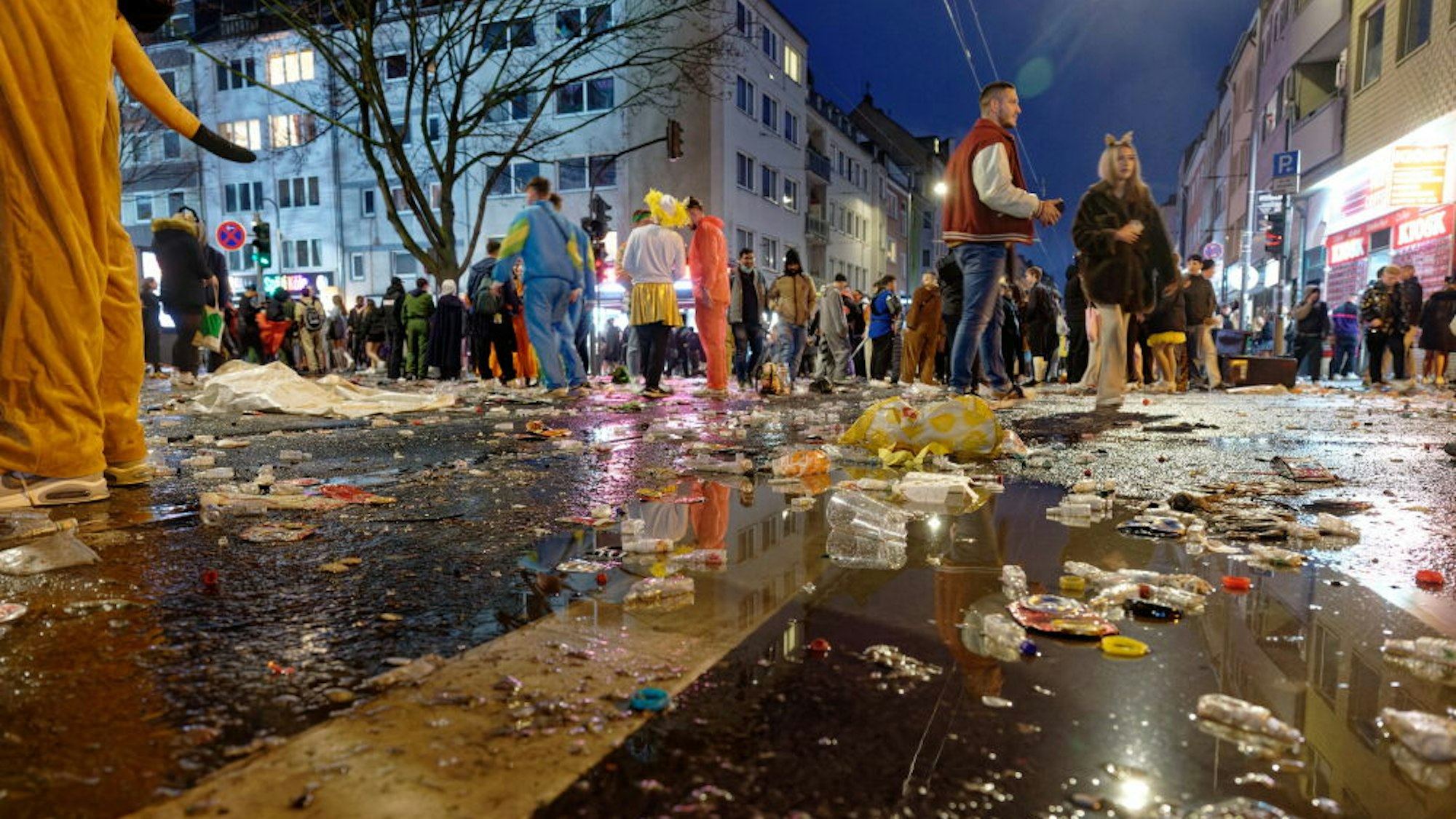 Müll liegt am späten Abend auf der Zülpicher Straße, im Hintergrund stehen kostümierte Menschen in kleinen Gruppen zusammen.
