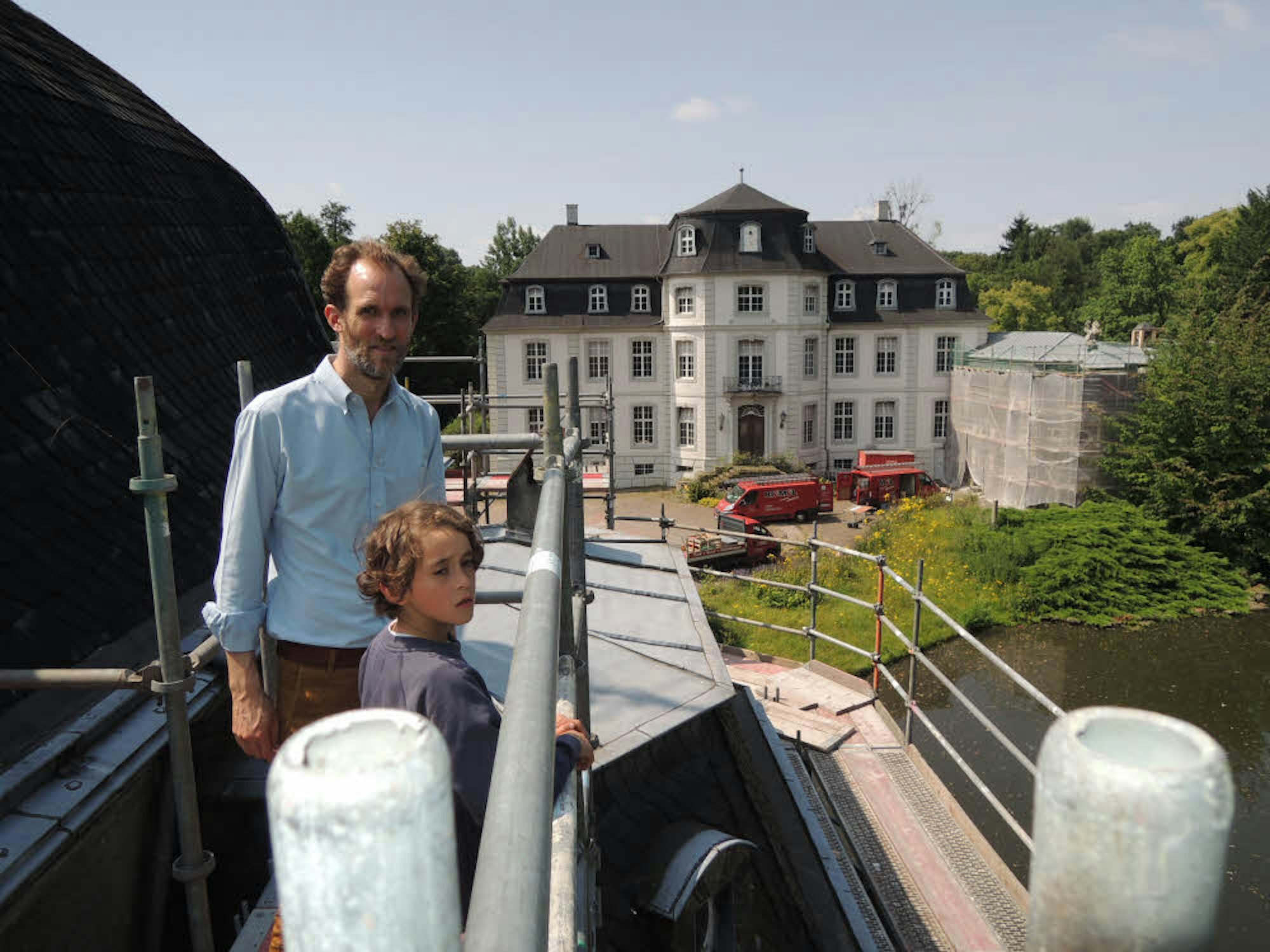 Severin Graf Hoensbroech mit Sohn Nepomuk blicken von einem Gerüst in den Park von Schloss Türnich. Am Dach der Kapelle neben dem Herrenhaus wird bereits gearbeitet.