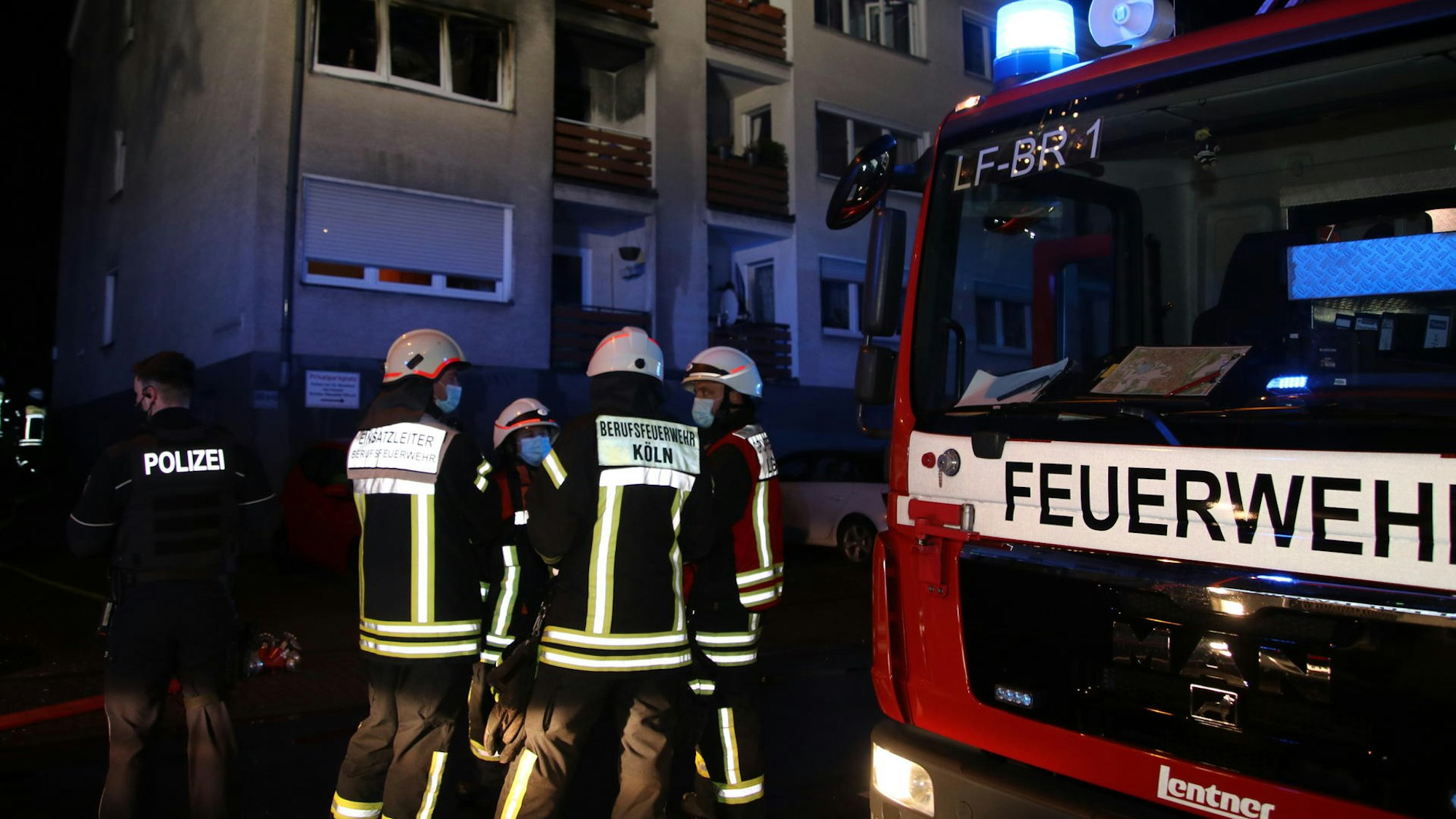Feuerwehrleute, ein Polizist sowie ein Löschfahrzeug stehen in der Nacht vor einem Mehrfamilienhaus.