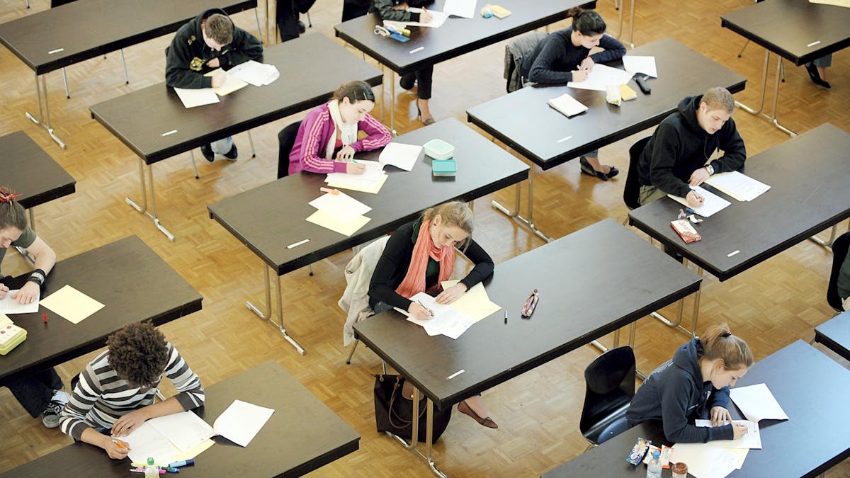 Junge Frauen und Männer sitzen bei einer Prüfung weit auseinander an Tischen.