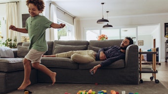 Ein Junge rennt durchs Wohnzimmer, während der Vater auf der Couch liegt.