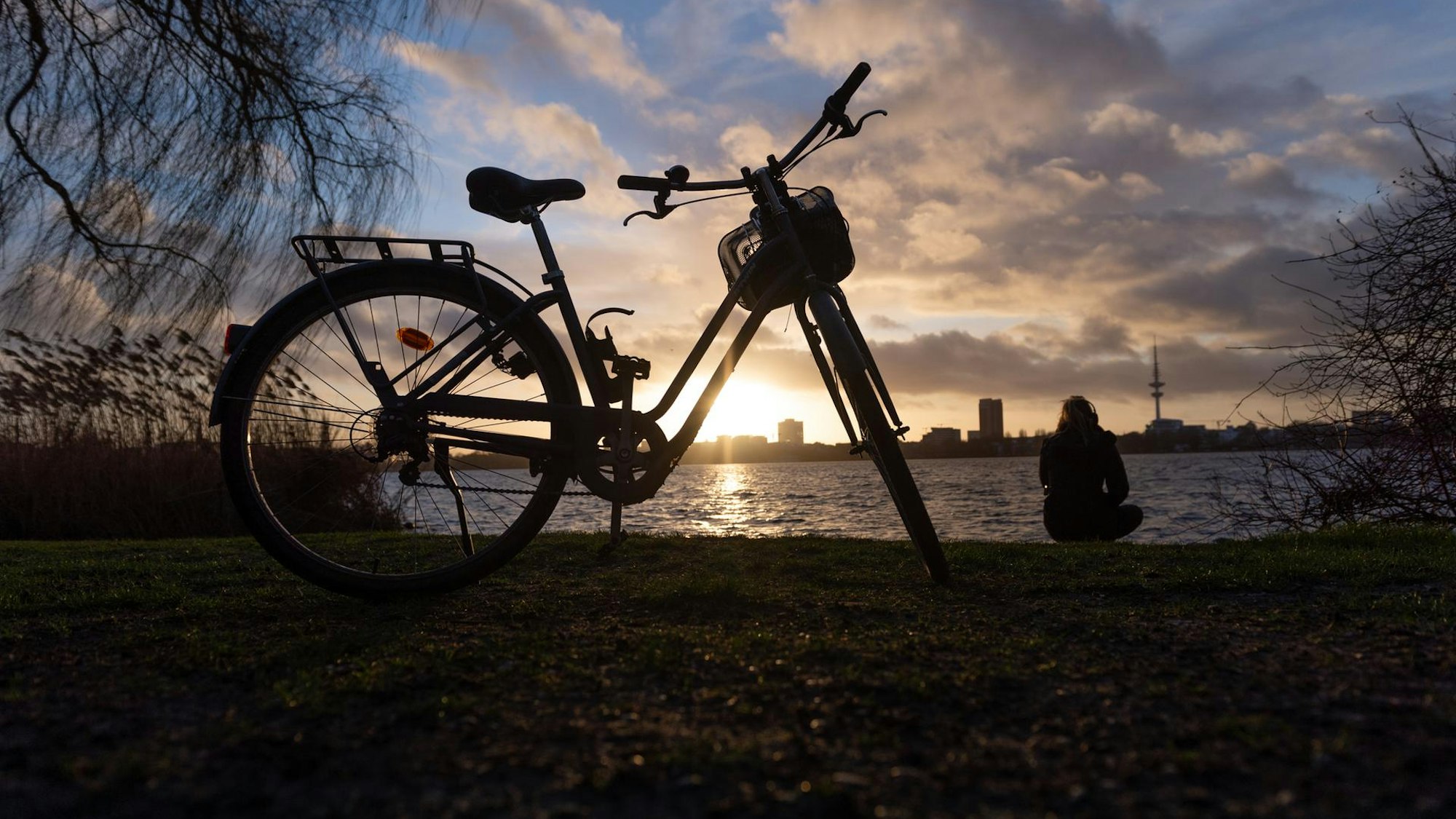 Ein Fahrrad steht am Rheinufer im Sonnenuntergang und eine Frau hockt am Wasser.