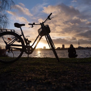 Ein Fahrrad steht am Rheinufer im Sonnenuntergang und eine Frau hockt am Wasser.