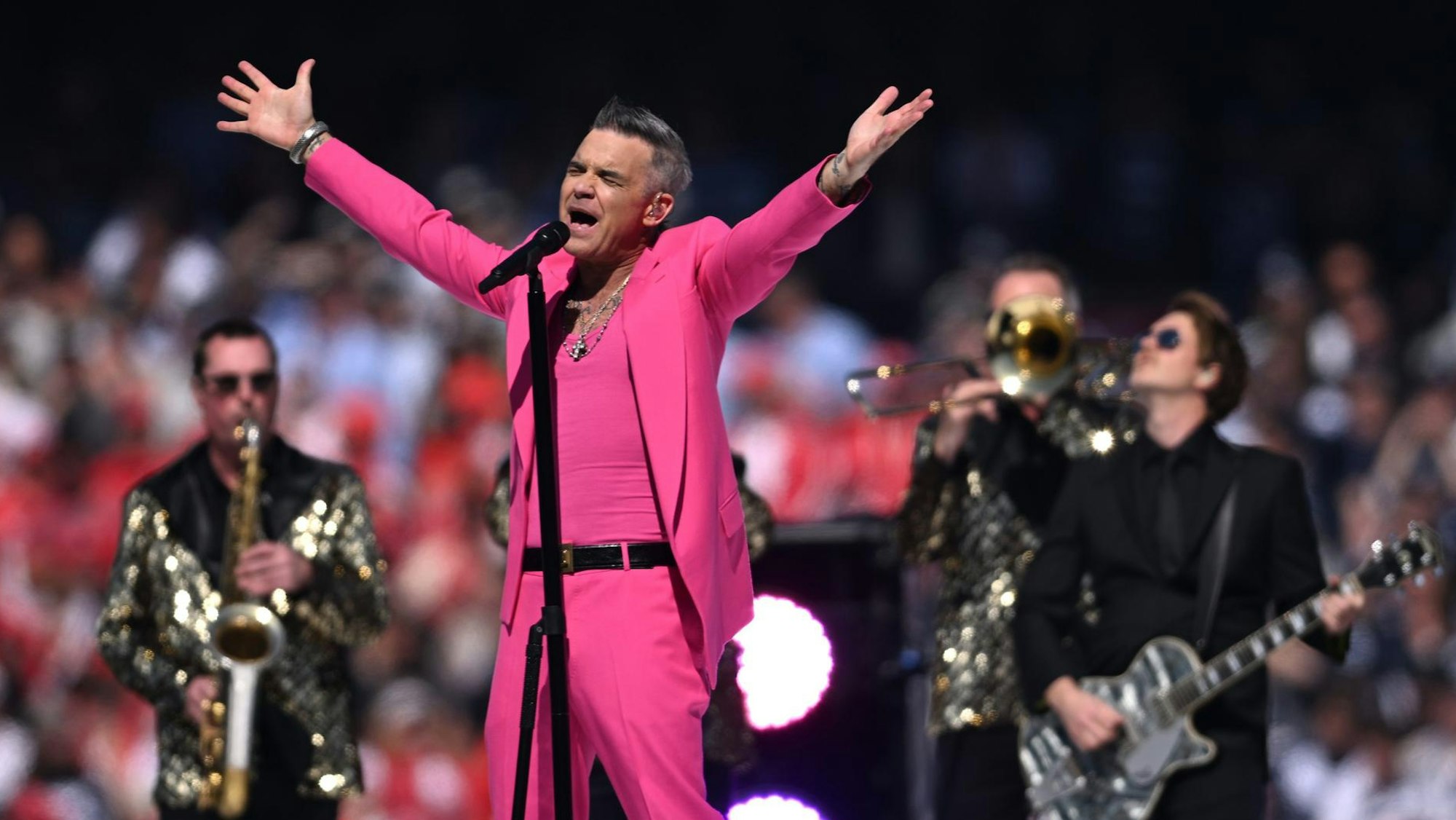 Robbie Williams streckt beim Live-Auftritt die Arme aus. Er trägt einen pinken Anzug und singt. Hinter ihm spielt seine Band mit Glitzer-Jacketts.