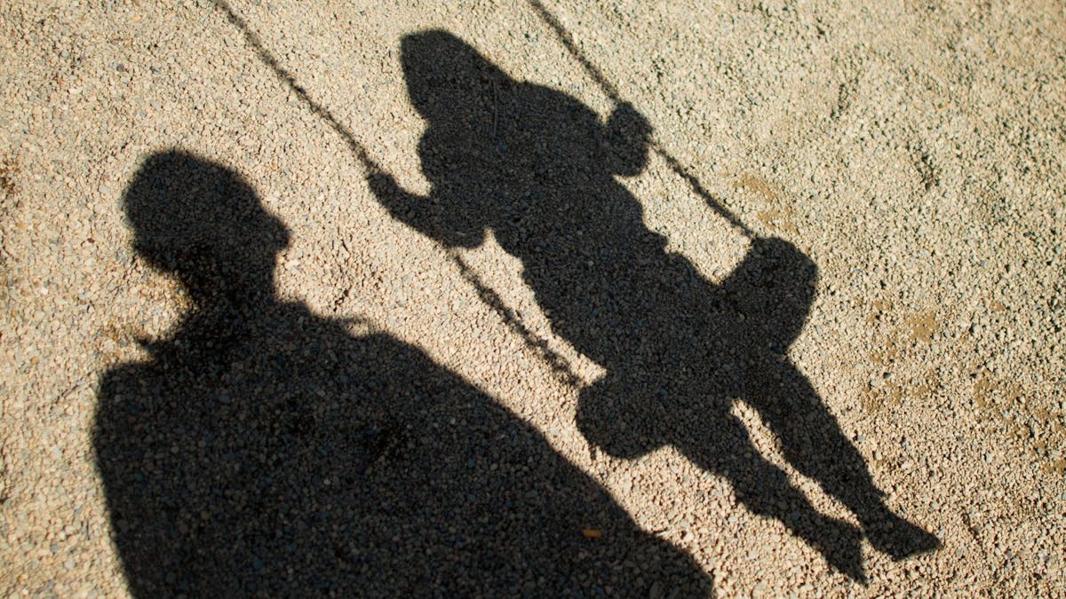 Der Schatten von einem Mann und einem schaukelnden Kind fallen auf Sand auf einem Spielplatz.