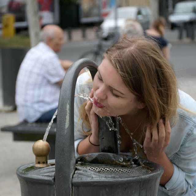 Das Foto zeigt eine Frau, die aus einem Trinkwasserbrunnen trinkt.