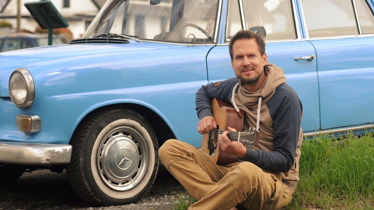 Mike Kremer von Miljö spielt sitzend Gitarre vor einem Auto