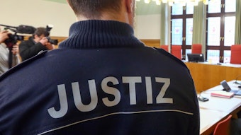 Ein Justizwachtmeister im Gericht. Auf der Rückseite der Jacke ist "Justiz" zu lesen, im Hintergrund sieht man Journalisten mit Mikrofon und Kamera.