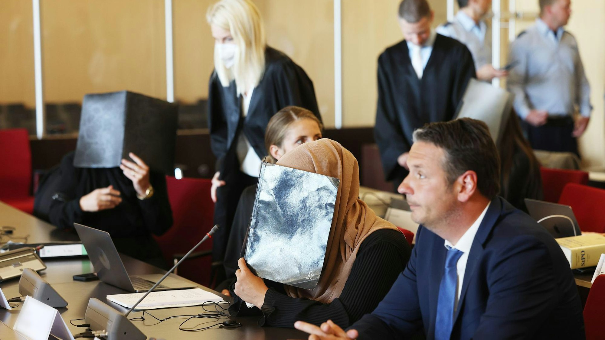 Ein Gerichtssaal: eine Frau mit Kopftuch hält sich eine Kladde vors Gesicht, daneben Anwälte.