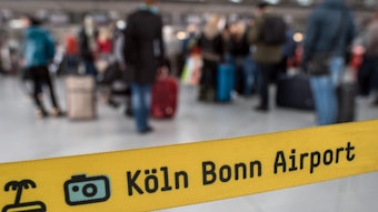 Hinter einem Absperrband, auf dem Köln Bonn Airport steht, stehen mehrere Personen.