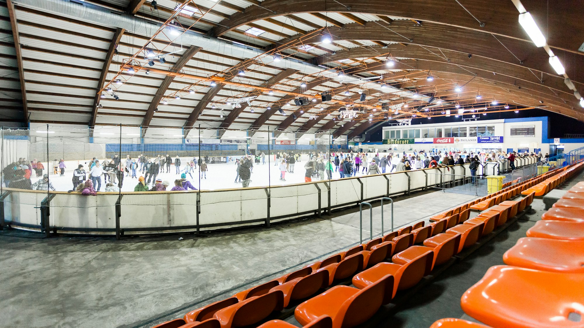 Innenraum einer Eissporthalle, Blick von orangen Sitzreihen auf die Eisfläche